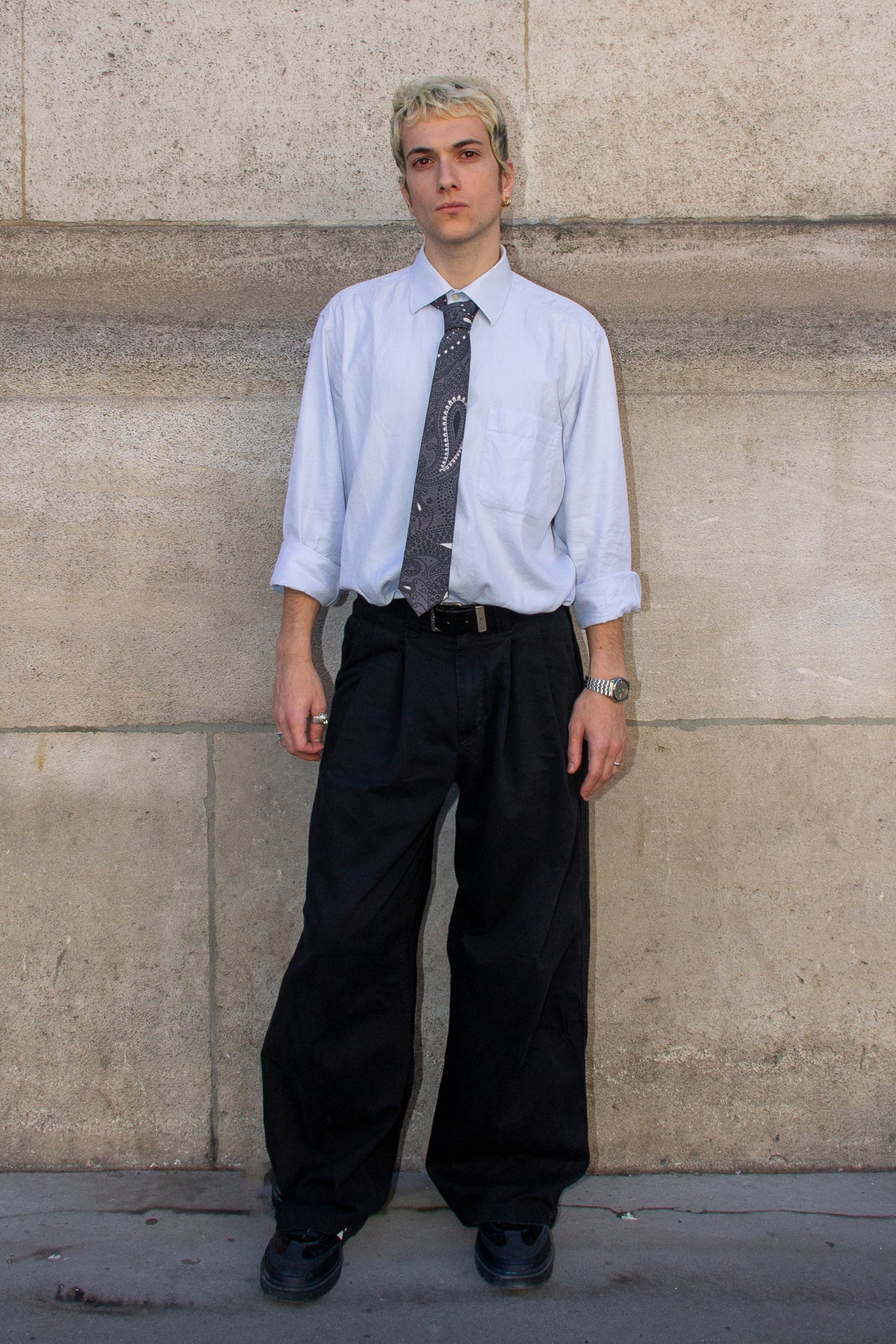 Homme vu de face portant une cravate à motifs paisley gris ardoise, une chemise bleu clair et un pantalon noir.