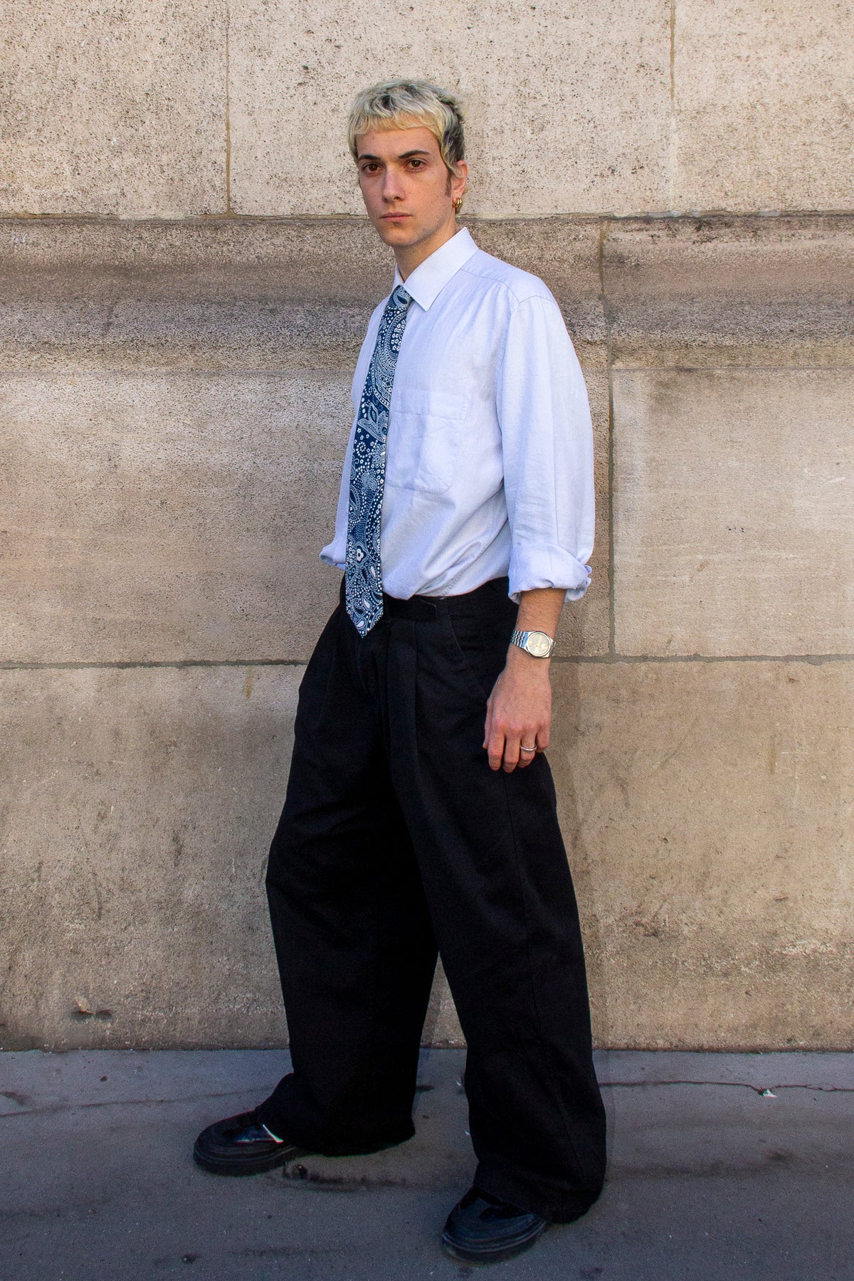 Un homme de 3/4 devant un mur qui porte la cravate en soie imprimée paysley bleu.