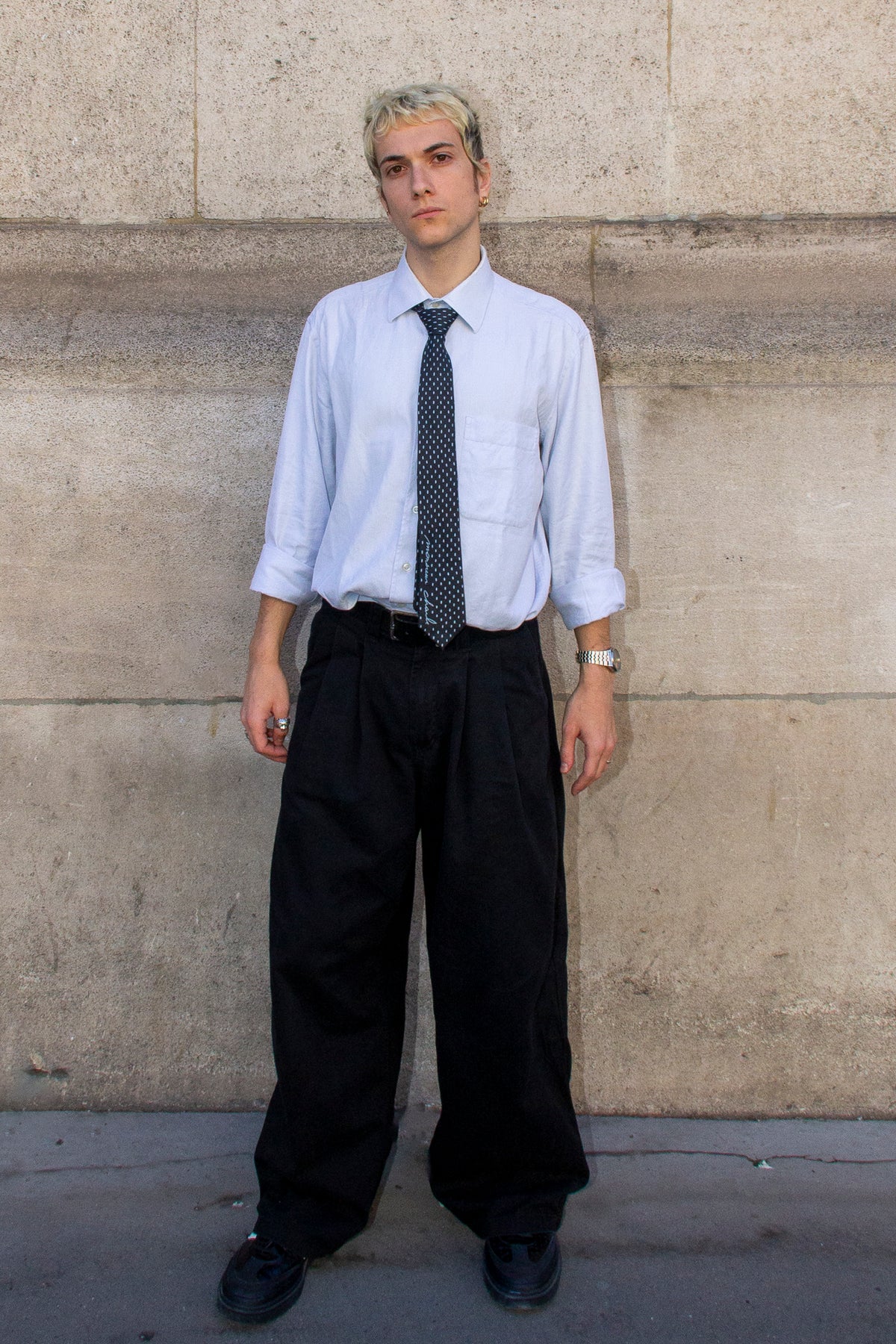 Homme vu de face portant une cravate noire à petits pois blancs, une chemise bleu clair et un pantalon noir.